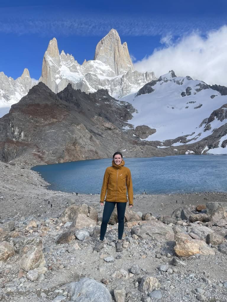 female in orange jacket at the summit of Laguna del Los Tres with mountain peaks in the background illuminated by the sunlight and a blue glacial lake below whille spending 2 weeks in Patagonia