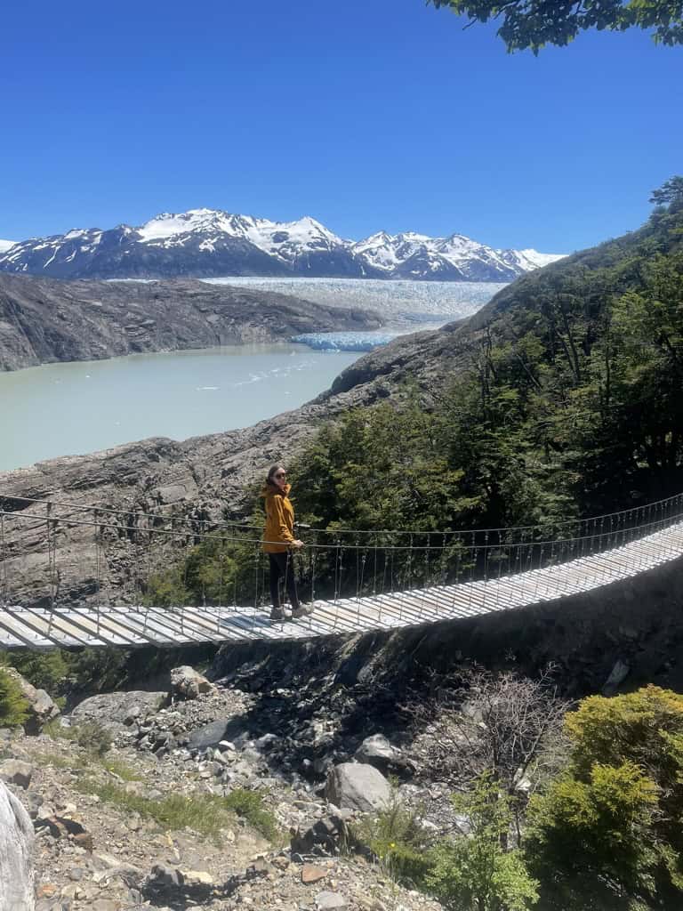 female in orange jacket crossing suspension bridge on a 2 week trip in Patagonia with view of Grey Glacier and grey lake below.