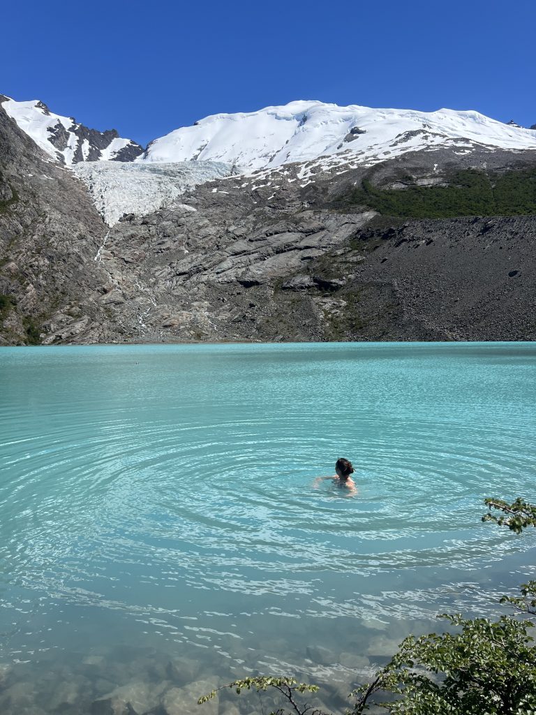 girl swimming in teal blue water of huemul galciar with a mountain range in the background