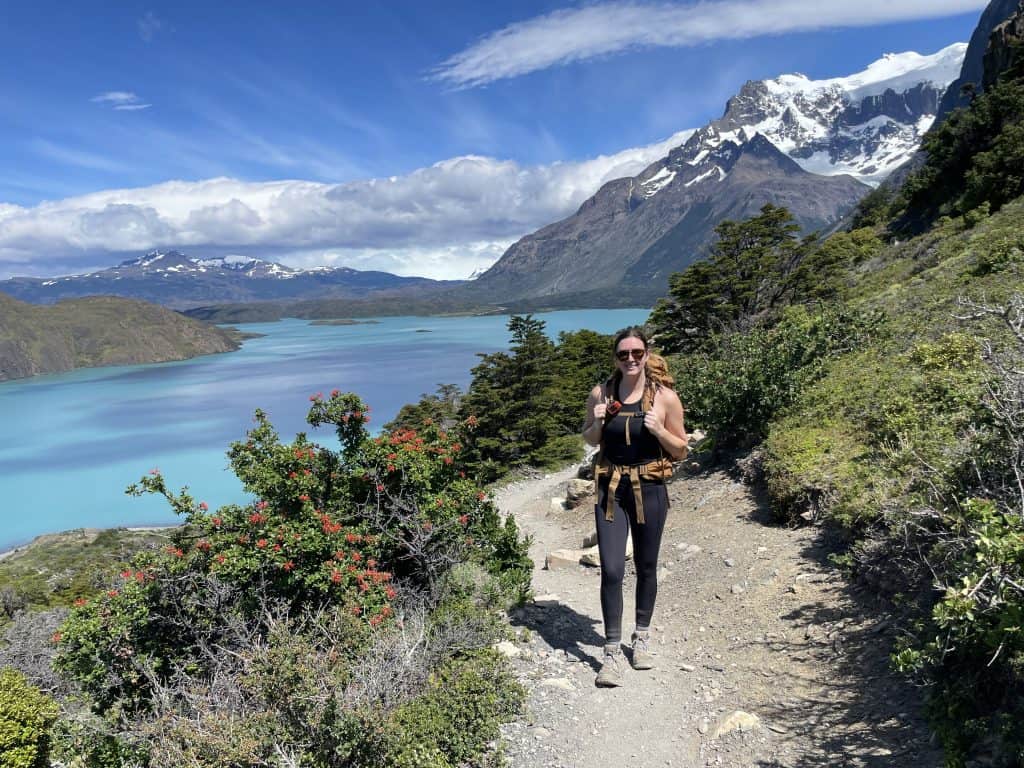hiker on dirt trail on the W-Trek trail on a two week trip in Patagonia with green bushes, bright blue lake and snow capped mountain tops