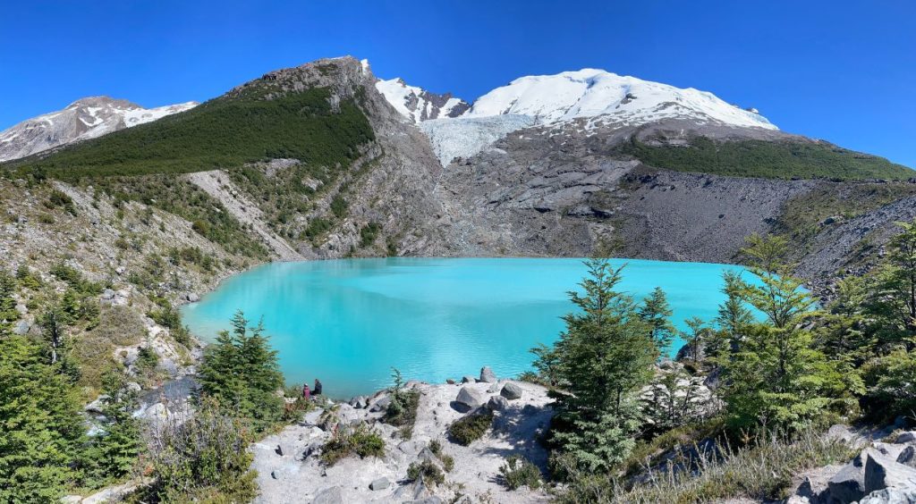 Huemul Glacial with bright teal coloured water, green trees and rocky snow capped mountain with a grey glaciar bleeding into the lake